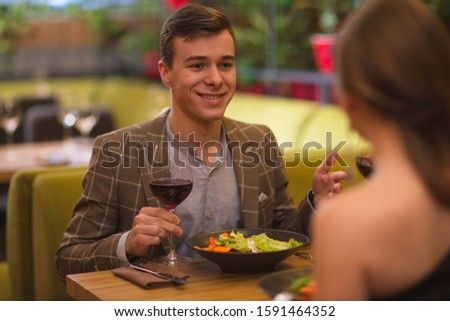 Handsome young man in checkered jacket dining in restaurant with wife. Happy man keeping glass of wine, talking and looking at woman sitting opposite. Concept of romantic atmosphere.