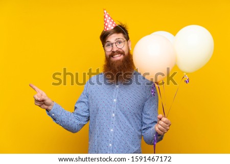Redhead man with long beard holding balloons over isolated yellow background pointing to the side to present a product