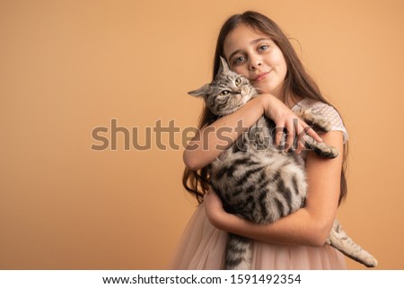 Beautiful cute brunette girl with loose hair, blue eyes and healthy skin hugging her grey cat and looking at the camera isolated over orange background, studio portrait