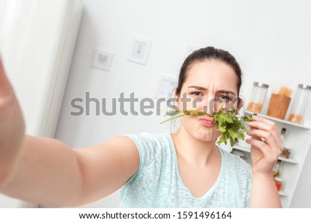 Young chubby woman standing in kitchen taking selfie picture on smartphone poing to camera making mustache with parsley grimacing playful close-up