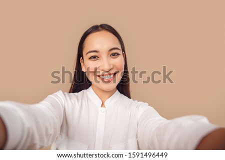 Young woman wearing white shirt with dark long hair standing isolated on bage background taking selfie photo on smartphone posing to camera smiling toothy close-up