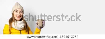 Young woman in a yellow jacket and hat holds a glass of coffee or tea on a light background. Emotion laughter, surprise, kiss. Concept winter, autumn, coffee house. Banner