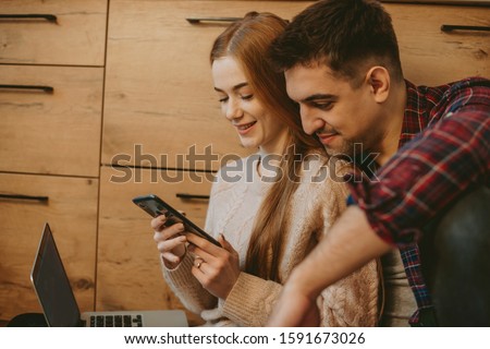 Side view portrait of a amazing couple sitting on the floor in the kitchen while red haired girl is holding a smartphone and her man is embracing from back