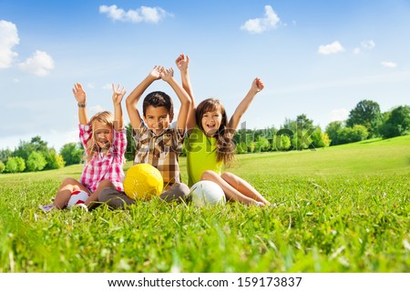 Portrait of three happy kids, boy and girls sitting in the grass in park with lifted hands and holding sport balls Royalty-Free Stock Photo #159173837