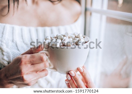 Girl in a white sweater holds a cup of hot chocolate with marshmallows and warms her hands staying near a window. Face is not visible.