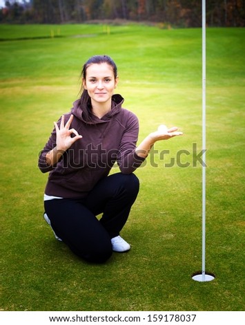 Smiling young woman golf player on green holding ball giving perfect shot sign