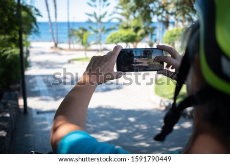 View from behind of a sporty woman with a bicycle helmet taking a photo with her cell phone. Horizon on the blue sea