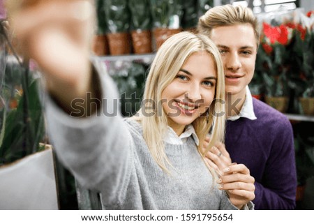 Beautiful young couple in casual clothes is making pictures of themselves with a flower and smiling while standing in store