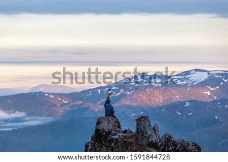 Panoramic mountain view. A clear sunny evening. Sunset. Beautiful blue clouds. At the top of the cliff sits a young man. A traveler controls a drone. Norway. Senja. Scandinavia.