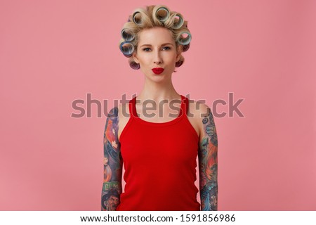 Portrait young attractive blonde woman with tattooes and festive makeup pouting her lips while looking positively at camera, having curlers on her head while standing over pink background