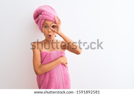 Beautiful child girl wearing shower towel after bath standing over isolated white background doing ok gesture shocked with surprised face, eye looking through fingers. Unbelieving expression.