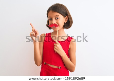Beautiful child girl holding fanny party mustache standing over isolated white background very happy pointing with hand and finger to the side