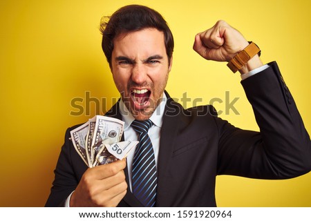 Young handsome businessman wearing suit holding dollars over isolated yellow background annoyed and frustrated shouting with anger, crazy and yelling with raised hand, anger concept
