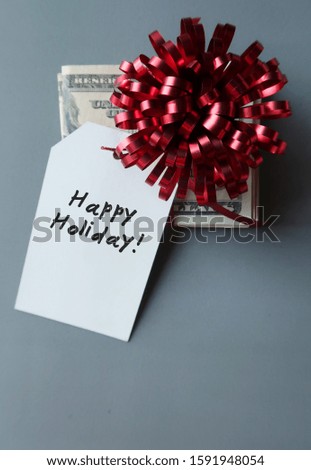 Cash Dollars money on gray background with red gift bow and a gift tag written HAPPY HOLIDAY! - concept of bonus year end or holiday gift money for employee or love ones