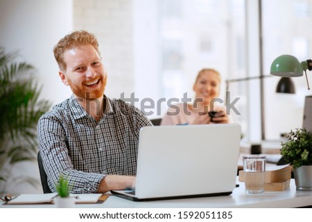 Young happy businessman using laptop at his office desk.  Casual man operating on his computer while his colleagues smiling in the background.
