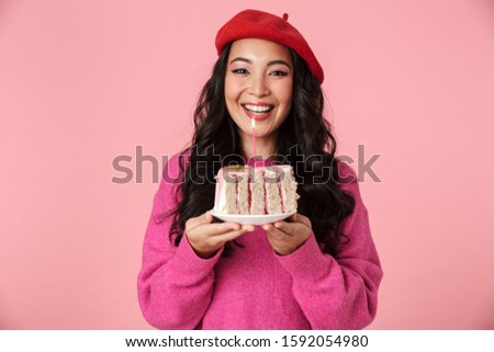 Image of excited beautiful asian girl smiling and holding birthday cake with candle isolated over pink background