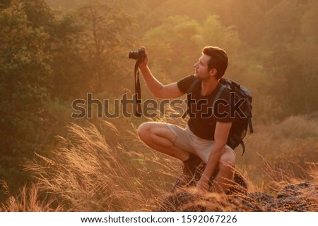 A man photographer and traveler holding digital camera taking photo of mountain with sunset background, gold lighting from sun 