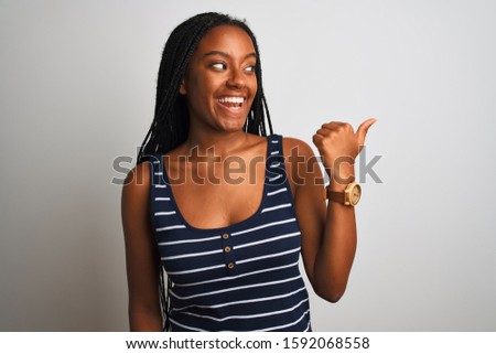 Young african american woman wearing striped t-shirt standing over isolated white background smiling with happy face looking and pointing to the side with thumb up.