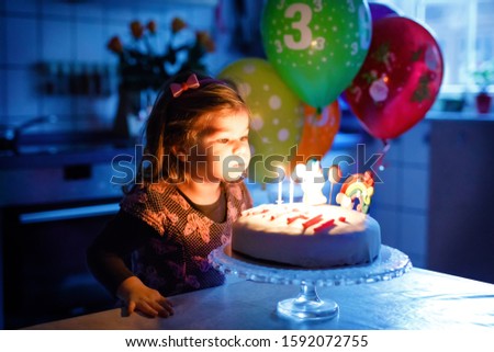Adorable little toddler girl celebrating third birthday. Baby toddler child with homemade unicorn cake, indoor. Happy healthy toddler is suprised about firework sparkler and blowing candles on cake