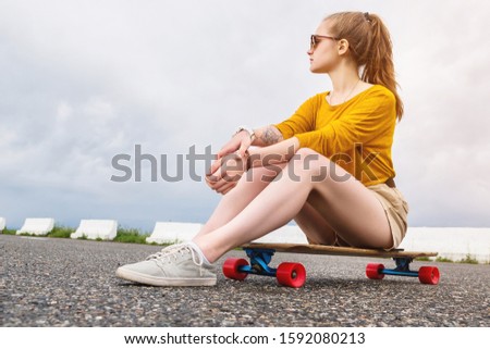 Attractive young girl with a tattoo on her arm in sunglasses and shorts sits on her longboard in a suburban parking on a background of gray sky. Millennials generation and leisure concept