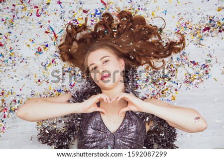 Portrait of tender cute pretty charming girl in a sparkling dress lying on the floor with carnival candy. Happy New Year 2020 celebration holiday concept. Showing shape heart with hands. Top view.