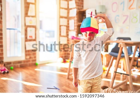 Beautiful blonde toddler wearing fanny colorful propeller cap. Standing around lots of toys at kindergarten