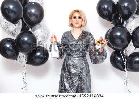 Cheerful girl in a silver dress at a party with champagne, confetti and black balloons against a white wall
