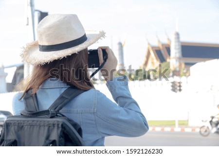 The back of a young tourist wearing a denim jacket, straw hat and a backpack standing to take pictures at the Phra Kaew Temple of Bangkok for holidays and travel.