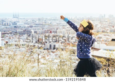funny little child girl posing powerful with her fist in the air disguised as a superhero with homemade costume in front of a cityscape, imagination and girl power concept