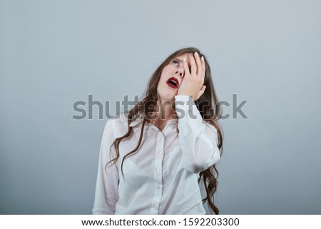 Bored caucasian brunette young woman in fashion white shirt keeping hand on eye isolated on gray background in studio. People sincere emotions, lifestyle concept.