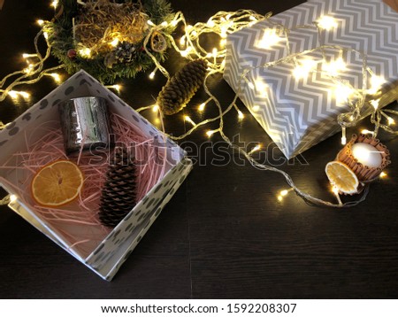 Silver gift box with shredded pink paper filled with candle, dries orange and pine cone, on background of Christmas garland and Christmas decorations on wooden table. Christmas and New Year presents. 