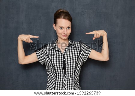 Studio half-length portrait of happy optimistic young woman wearing shirt, smiling cheerfully, pointing with index finger at herself, being proud of her deeds or results, isolated over gray background