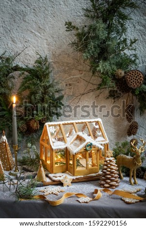 Gingerbread house stands on a wooden table among gingerbread toys. Everything is strewn with snow. Candle is burning nearby. On the background is a wreathes of pine branches and cones. Close view.