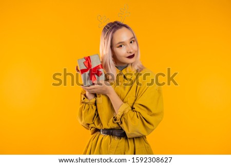 Unusual woman with pink hair smiling and holding gift box on yellow studio background. Hipster girls portrait in trendy outfit. Christmas mood.