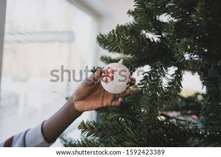 Closeup image of colored girl holding white holiday bauble with red snowflake on it hanging on Christmas tree.