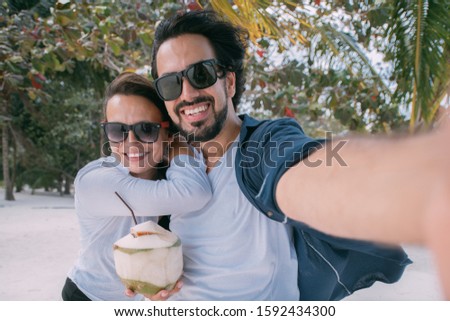 A pair of lovers take a selfie under a palm tree on a tropical beach. A guy and a girl are photographed among tropical plants and palm trees. Newlyweds Enjoy Tropical White Sand Lagoon
