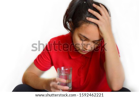 A young girl on a white background of European appearance in a red Polo with dark hair gathered in a bun closed her eyes and holds one hand over her forehead, the other hand holding a glass of water, 