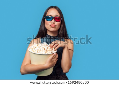 Young woman with dark long hair wearing red-blue 3d glasses standing isolated on blue background holding bucket eating popcorn pouting lips posing to camera playful
