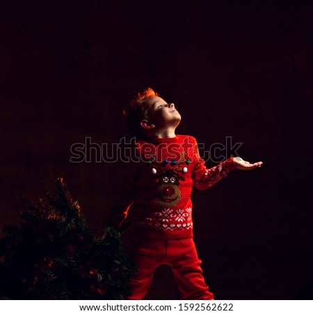 Boy kid catches snowflakes with his palm holding in one hand a Christmas tree. The child is dressed in a knitted suit with a sweater with a picture of a deer