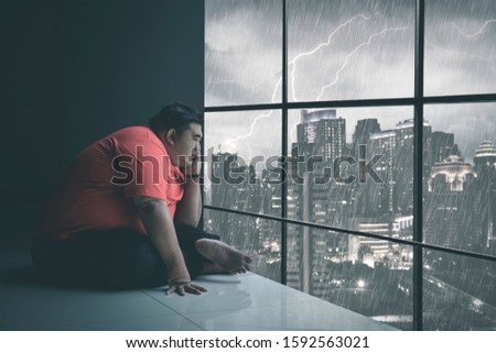 Side view of Asian man holding his chin while staring at the raining city through the window somberly with raining cityscape background