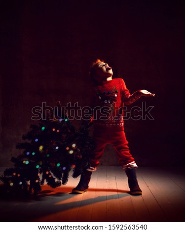 Boy kid catches snowflakes with his palm holding in one hand a Christmas tree. The child is dressed in a knitted suit with a sweater with a picture of a deer