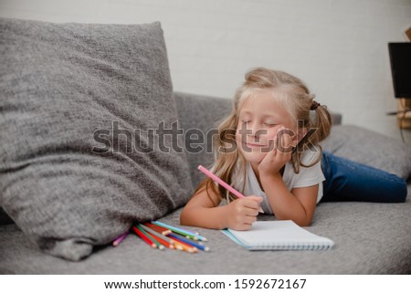 Little girl drawing lying on thesofa in the living-room. White abstract wall in the background. Horizontal view drawing and dreaming concept.