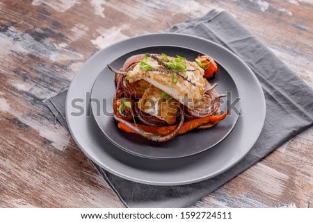 Appetizing cod filet with red onion and herbs: sage, rosemary and thyme, baked potatoes and carrots on a side dish in a gray plate on a gray napkin. Tasty and healthy sea fish