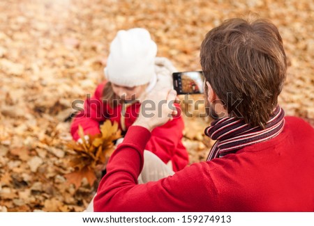 Man taking autumn outdoor picture with mobile phone. Father photograph his girl child playing in the park.