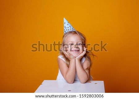 Portrait of a little blonde girl in a festive hat on a colored background and confetti.