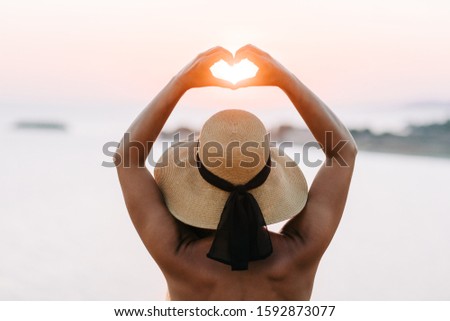 Young woman making a heart shape into the sunset by the sea in sunset