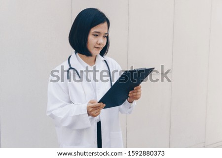 an attractive Asian female doctor in white coat with a black stethoscope, holding a black clipboard, white wall background
