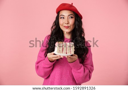 Image of excited beautiful asian girl smiling and holding birthday cake with candle isolated over pink background