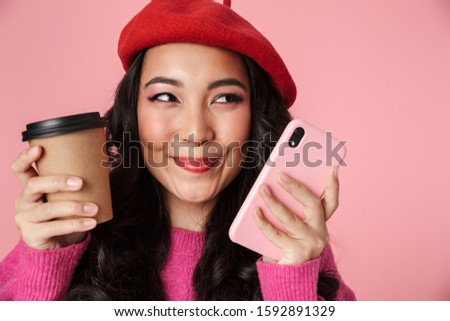 Image of young beautiful asian girl with long dark hair wearing beret holding smartphone and coffee cup isolated over pink background