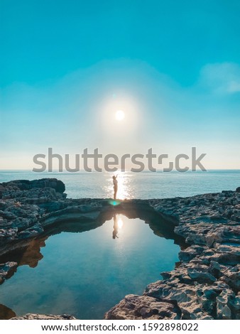 Sunset at Blue Window on Gozo Island in Malta. In the picture is a girl with her hand up holding the sun overlooking the horizon.